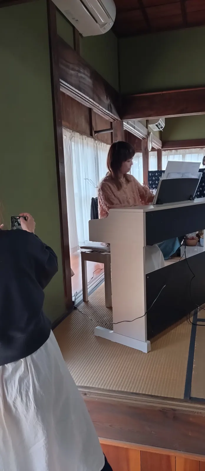 白い電子ピアノの譜面を見ながら演奏する女性。窓からの光の中、横顔がはっきり見える
