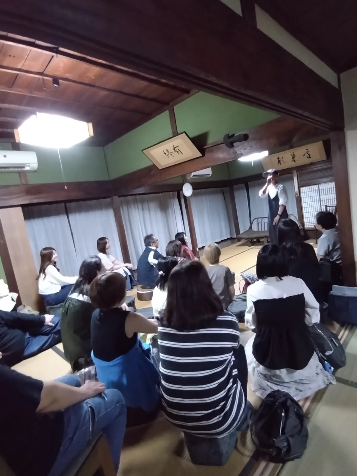 夜、和室で座って舞台を見つめる観客の後ろ姿。奥に小さく演者が立つ