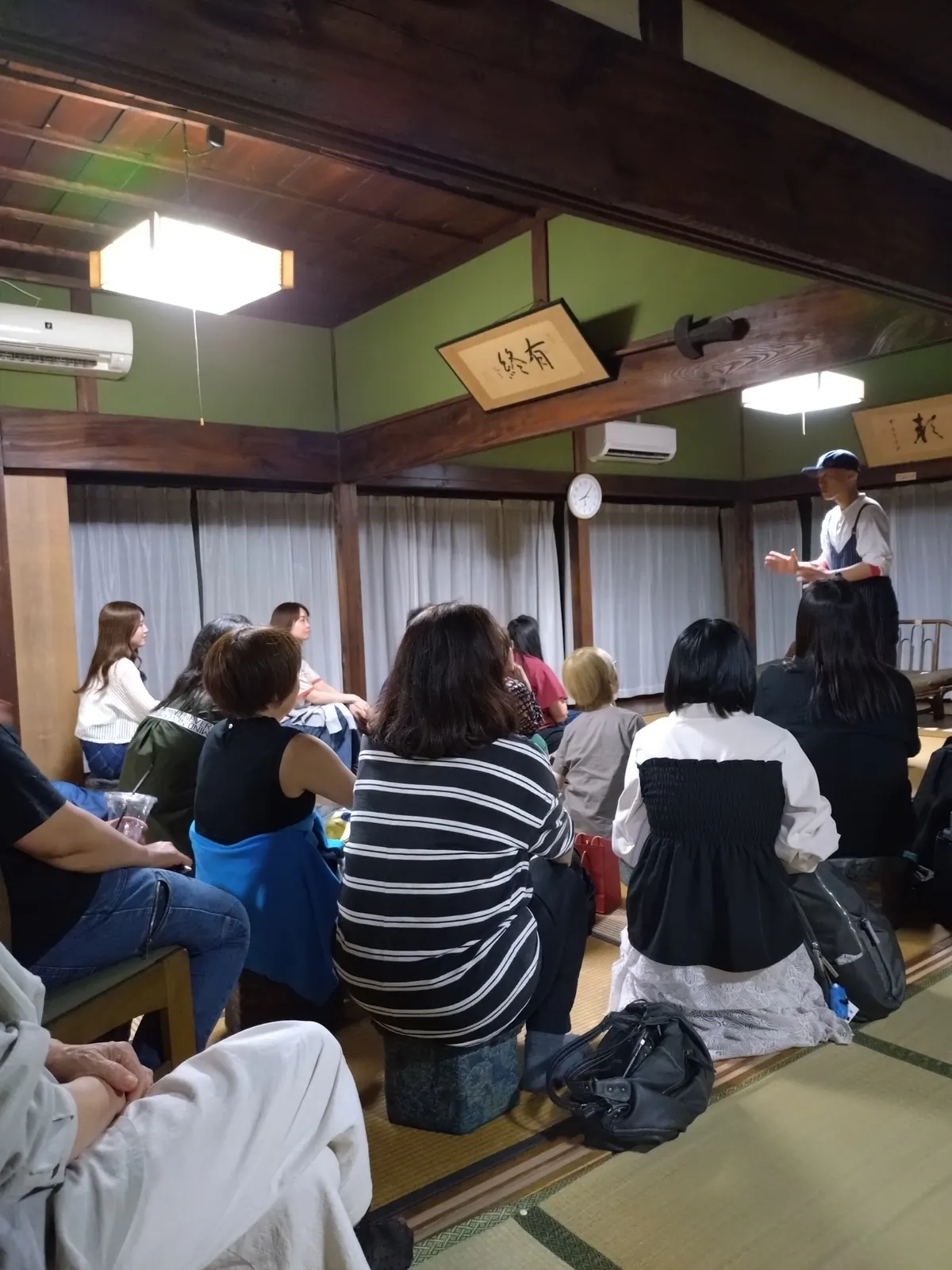 うす暗い和室。台の前に立つ演者と、見つめる人たち